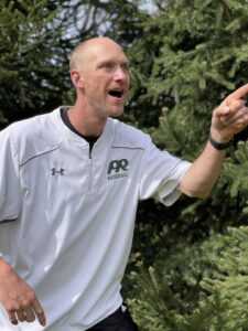 Bald coach in white jersey pointing and motivating outdoors, green blur background.